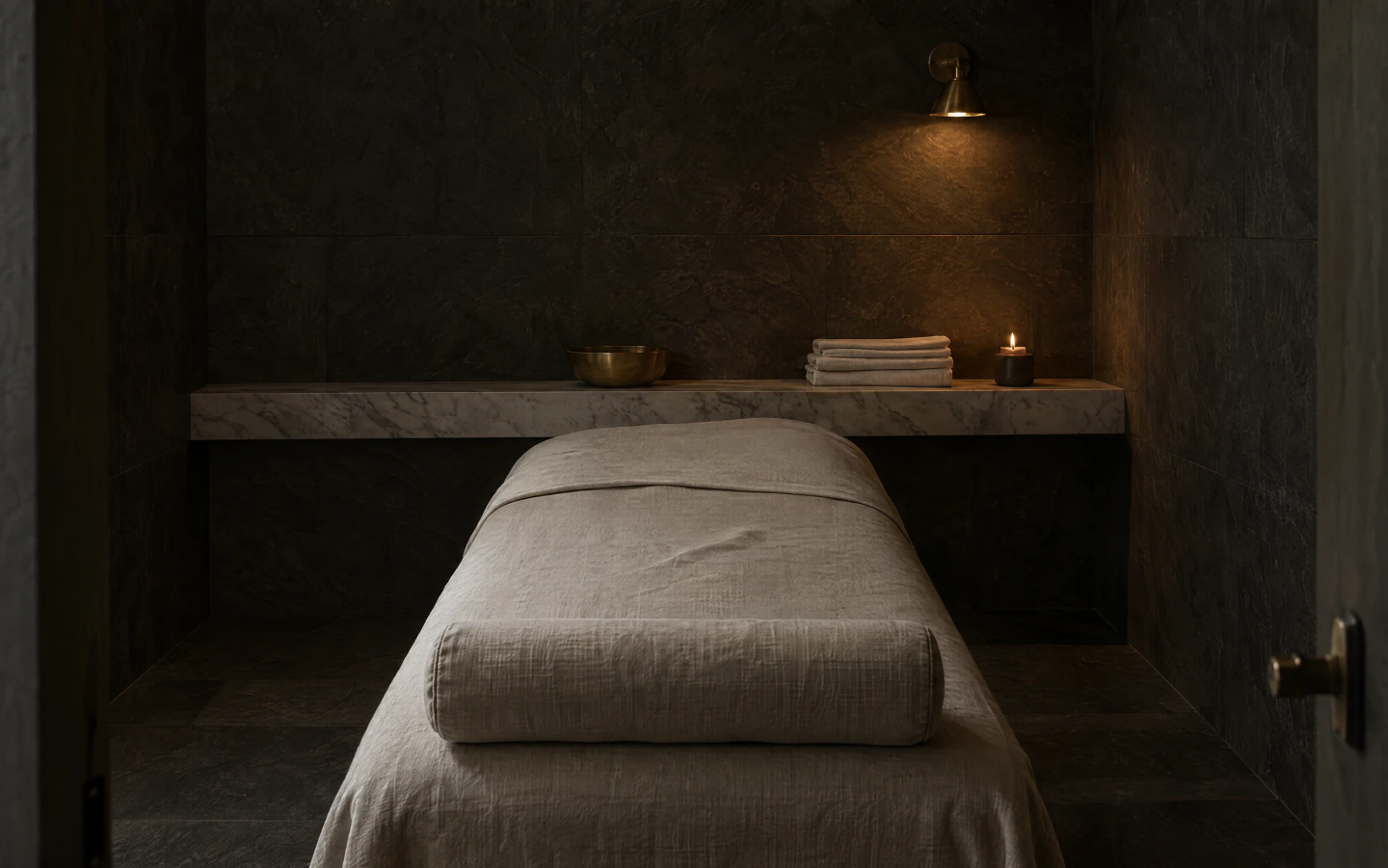 A treatment room in dark honed stone, a bed dressed in cream linen under a marble ledge with a lit brass sconce above it.