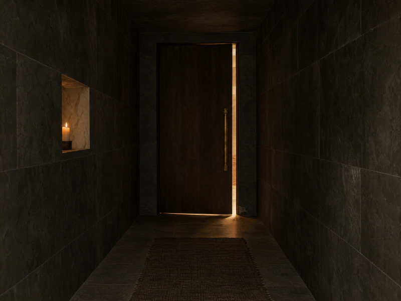 A tall dark timber door with a long aged brass handle at the end of a charcoal stone hall, warm light spilling from the room beyond.