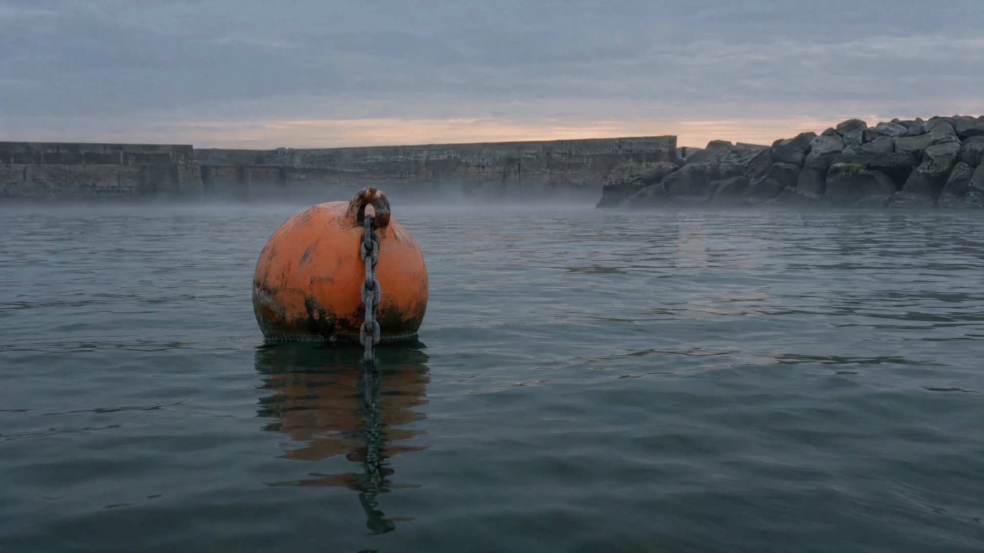 An orange mooring buoy on flat grey water at first light, its chain running down into the harbour, a stone breakwater behind.